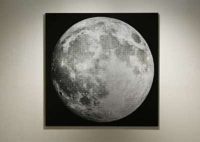 A textured artwork of the moon displayed on a square canvas against a plain wall, capturing detailed lunar features in monochrome tones.