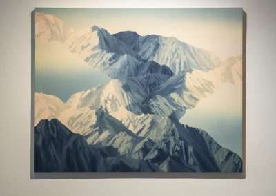 A framed, polygonal style art piece of a mountain range, displayed on a white gallery wall, with prominent white and blue hues creating a striking visual contrast.