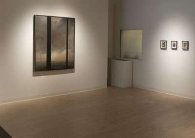 A clean, well-lit art gallery with wood flooring, displaying various framed artworks on neutral colored walls. One large painting dominates the left wall, with smaller pieces arranged on adjacent walls.