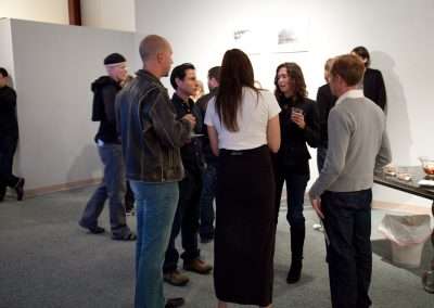 A group of people engaging in conversation at an art gallery event, with artwork displayed on the walls and refreshments on a nearby cart.