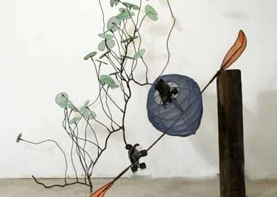 Sculpture depicting a stylized plant with round, blue petaled flowers and green leaves, created with wire and attached to a wooden base, incorporating abstract, minimalist elements.