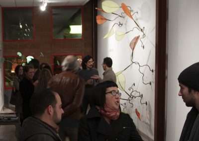 People gather at an art gallery opening, engaging in conversation near a wall adorned with a large, leafy vine mural. The setting is warmly lit with a casual atmosphere.