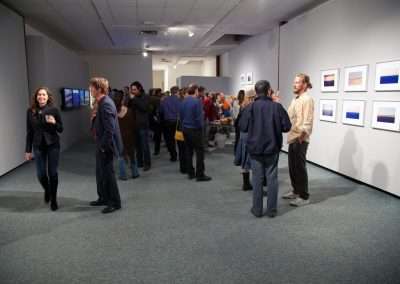 A lively art gallery opening with people standing and conversing. The room has white walls decorated with framed pictures of varying blue shades.