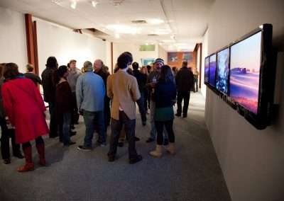 A group of people viewing digital artworks displayed on wall-mounted screens in a brightly lit gallery, engaging in conversation and observing the art.