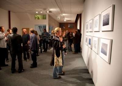 People viewing photographs at an art gallery exhibition, engaging in conversations and observing various displayed artworks on the walls.