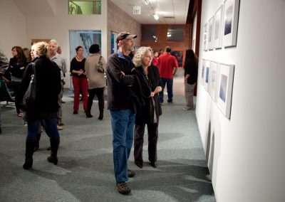 People observing framed artwork on a white wall in a gallery. A man in a baseball cap and a woman with curly hair stand closely viewing one exhibit. Other visitors are dispersed, engaging with various displays. The gallery has a bright, modern interior.