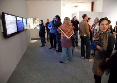 People viewing art at a gallery exhibition, with some standing and talking, while others are observing paintings displayed on the wall-mounted screens.
