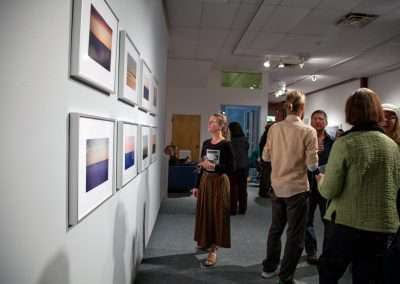 Guests interact and view artwork at a gallery opening, featuring framed landscape paintings on white walls in a well-lit indoor setting.