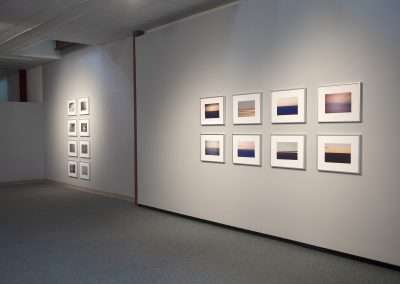 A serene art gallery hallway with two white walls displaying sets of framed photographs, featuring various light and landscape themes. Soft lighting from the ceiling illuminates the space.