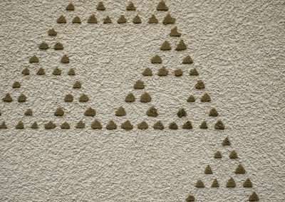 A beige textured wall features a design created with small, raised triangular shapes arranged to form larger triangles, creating a geometric, fractal pattern. The triangles vary in size, with smaller triangles clustering to form the edges of larger ones.