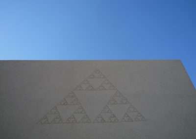 A large triangular fractal pattern etched on a concrete wall under a clear blue sky.