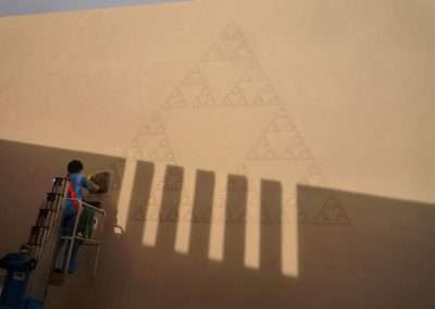 A worker on a lift paints a large geometric mural of nested triangles on a building's exterior wall, casting sharp shadows on the surface during a sunny day.