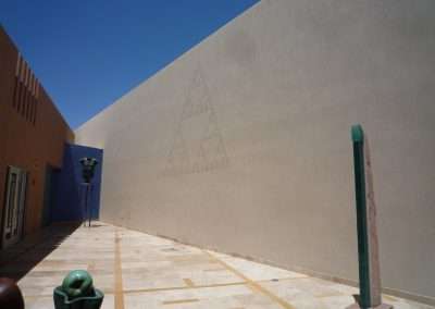 A spacious outdoor courtyard featuring a large plain wall with a geometric triangular pattern, accompanied by sculptures, including a blue head and a green pillar, under a clear sky.