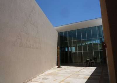 Modern architectural building with a large glass entrance, visible under a clear blue sky, flanked by a beige wall with a geometric relief pattern on the left.