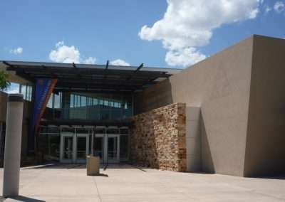 A modern building entrance with glass doors, featuring distinct architecture with a stone wall section and geometrically shaped concrete walls under a clear blue sky.
