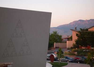 A large concrete wall with a triangle pattern on the left and a scenic view of a mountain range bathed in the glow of sunset in the background. Partial view of a building and parked cars are visible on the right.