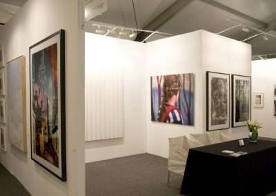 An art gallery space with white walls displaying various large paintings and photographs, including abstract and figurative works, with a table and flowers in the foreground.