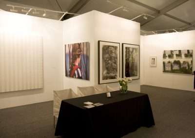 An art gallery interior featuring a modern exhibition space with a variety of framed artworks on white partition walls, a black table with brochures and a vase of flowers in the foreground, and a person standing to the right.