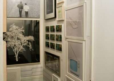 An art exhibition corner displaying various framed artworks on white walls, including abstract designs, a photograph of two people, and a shelf with small colorful items.