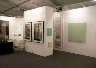 An art gallery exhibition with various artworks displayed on white panels, including photographs and abstract paintings, under soft lighting. Empty chairs add to the serene ambiance.