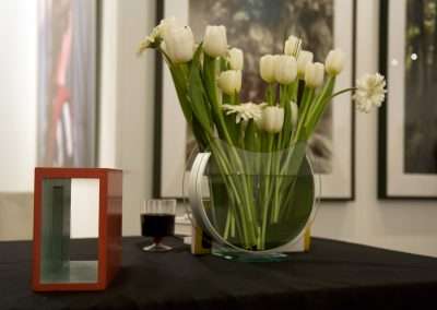 A vase of white tulips on a black table, accompanied by a red picture frame, a glass of red wine, and blurred art on the walls in the background.