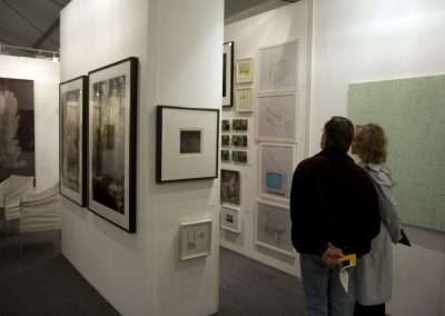 Two people view various framed artworks displayed on white walls in a well-lit gallery setting.