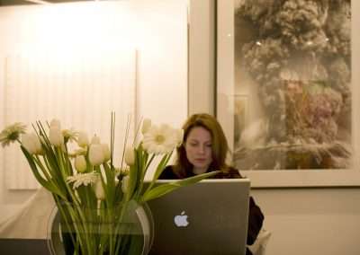A woman working on a MacBook in an art gallery, with fresh white flowers in the foreground and a large framed artwork in the background.