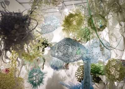 Art installation featuring an array of colorful, translucent sculptures resembling underwater life forms, crafted from recycled materials, with light casting intricate shadows on the surroundings.