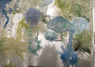 An art installation by Aurora Robson featuring an array of delicate, translucent sculptures made from recycled materials that resemble underwater sea creatures such as jellyfish, suspended from the ceiling in a gallery space with white walls.