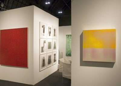 An art gallery corner displaying various artworks: a large red textured canvas on the left, a series of framed pieces on a white wall, and a yellow gradient canvas on the right.