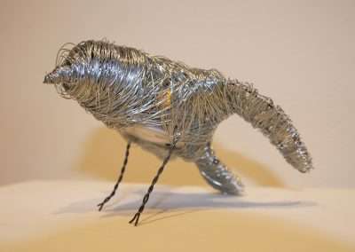 A sculpture of a dinosaur made from twisted silver wire, featuring detailed body, legs, and tail, displayed against a plain, light-colored background.