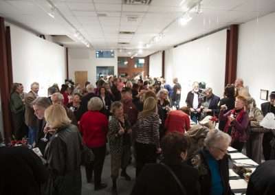 A bustling indoor art exhibition with a diverse group of adults mingling and viewing various artworks displayed on the walls.