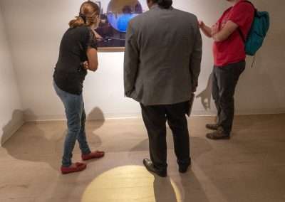 Three people observe an artwork with a golden circle displayed on a monitor in a gallery. Two men and one woman are engaged in discussion about the piece.