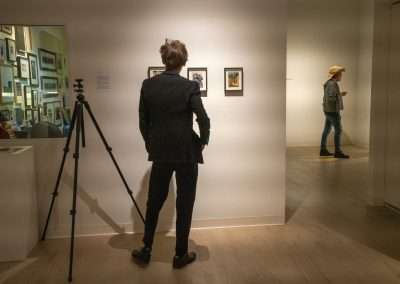 A man in a black suit examines artwork in a gallery, a tripod camera next to him, while another person in a beige hat observes art in the background.