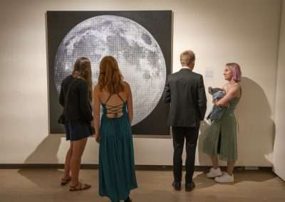 Three people view a large photograph of the moon in a gallery, with one person wearing a suit and the other two in dresses, one with purple hair.