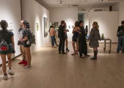 Visitors view diverse art pieces at a gallery exhibition, engaging in discussions and observations across a warmly lit room adorned with a variety of framed artworks.