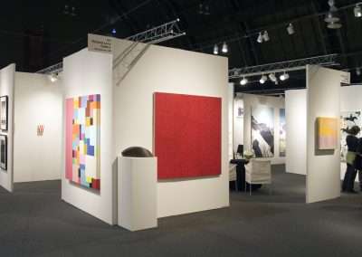 An art gallery display featuring various abstract paintings, including a prominent red canvas, and geometric shapes on white partitions under gallery lighting.