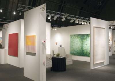 An art exhibition with several large abstract paintings in shades of red and green displayed on white partition walls, under bright lighting. A small seating area is in the center.