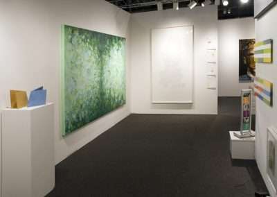 A contemporary art gallery with various styles of artwork including paintings and sculptures displayed on white walls and pedestals. The room features a large, leafy green painting as a focal point.