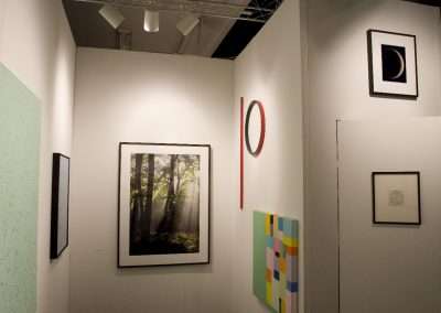 An art gallery at PULSE New York showcases various framed artworks on white walls, including a photograph of sunlit trees, an abstract geometric painting, and a black and white image of a partial circle. A red circular art piece is displayed prominently on a corner wall.