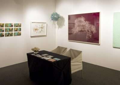 Art gallery exhibition room in New York featuring diverse paintings on the walls and a central table displaying books, with an empty chair nearby.