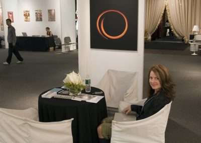 A woman smiles while sitting at a small round table with a white tablecloth and flowers, in a stylish art exhibition area with paintings and elegant drapes. A glowing orange circular artwork hangs on a nearby column.
