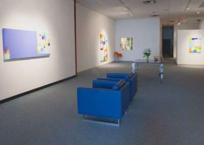 A modern art gallery with bright, abstract paintings displayed on white walls, blue benches in the foreground, and soft lighting illuminating the space.