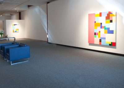 A modern art gallery with colorful geometric abstract paintings displayed on white walls, blue sofas in the foreground, and soft lighting from above.