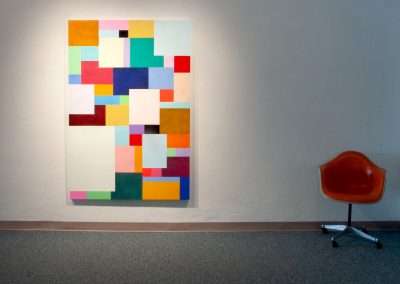 A modern abstract painting featuring a mosaic of colorful squares and rectangles on a large canvas, displayed beside an orange chair on a gray floor against a white wall.