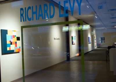 View through a glass door of the Richard Levy Gallery, displaying colorful abstract art pieces on well-lit white walls inside. The gallery name is printed on the glass door in bold letters.