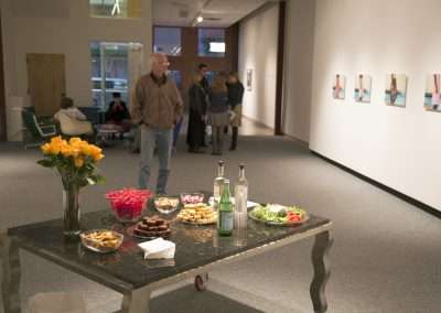 Art gallery interior featuring visitors viewing artwork, with a refreshment table in the foreground displaying an assortment of snacks and a vase of flowers.