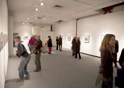 People viewing various framed photographs displayed on white walls in a well-lit art gallery. Several visitors are scattered around the room, some closely examining the artworks.