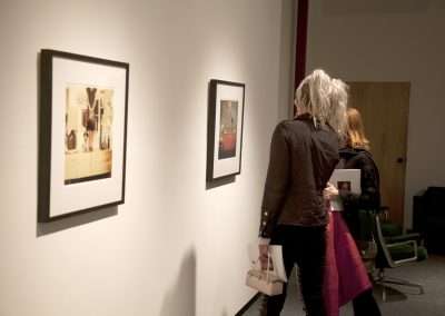 Two people observing framed photographs on a gallery wall, one individual is pointing at a picture while the other looks on. They carry bags and stand in a warmly lit indoor space.