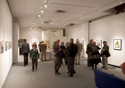 People viewing various artworks in an art gallery with white walls and soft lighting. Some visitors are standing and looking at the pictures, while others are engaged in conversation.